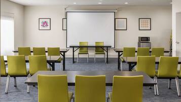 A conference room with a projector screen, green chairs, tables, and a microwave on the wall.