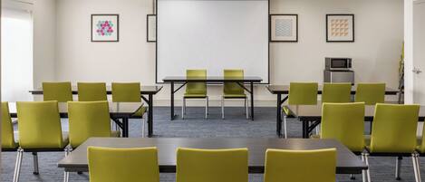 A conference room with a projector screen, green chairs, tables, and a microwave on the wall.