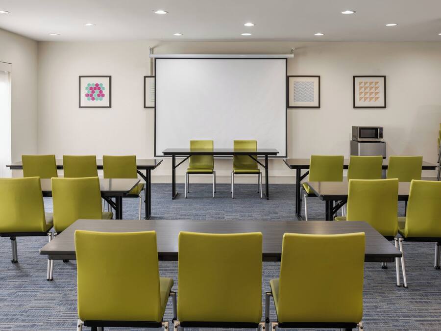 Un salón de conferencias con una pantalla de proyector, sillas verdes, mesas y un microondas en la pared.