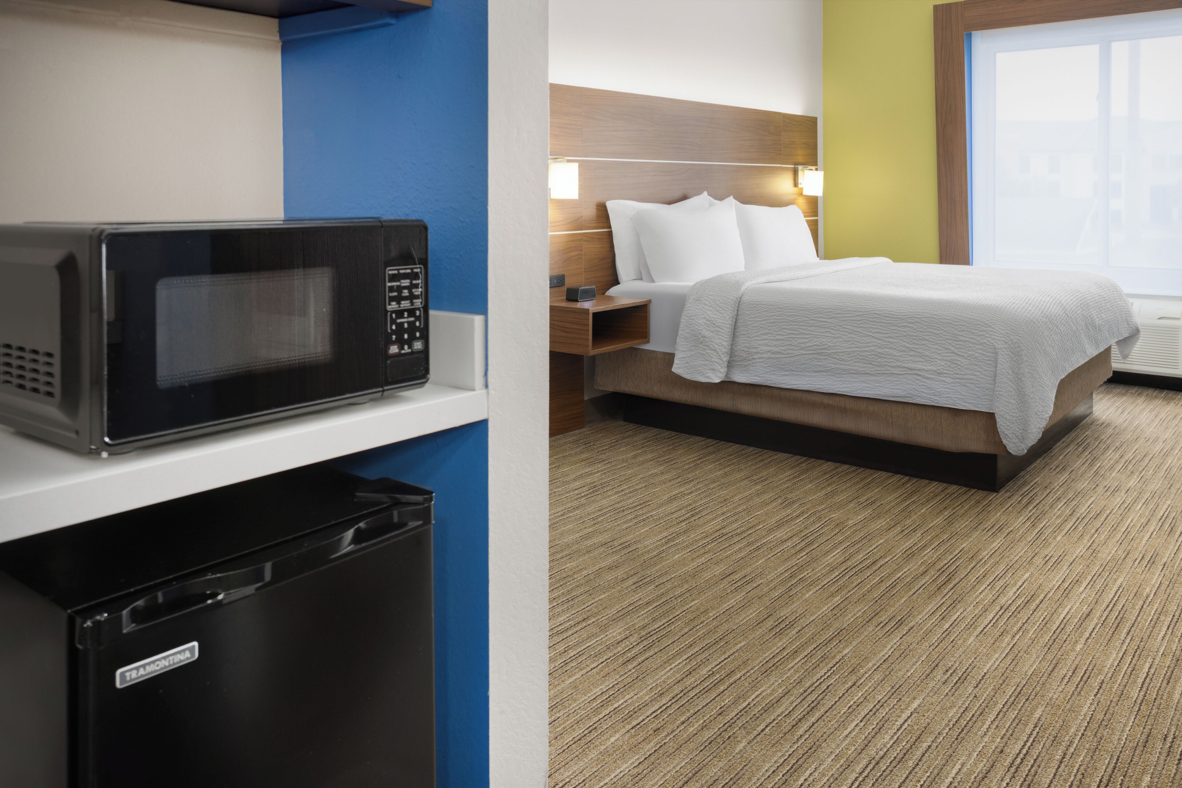 Habitación de hotel con microondas, un cesto de basura pequeño, cama con ropa blanca, mesita de noche con lámpara y ventana con vistas.