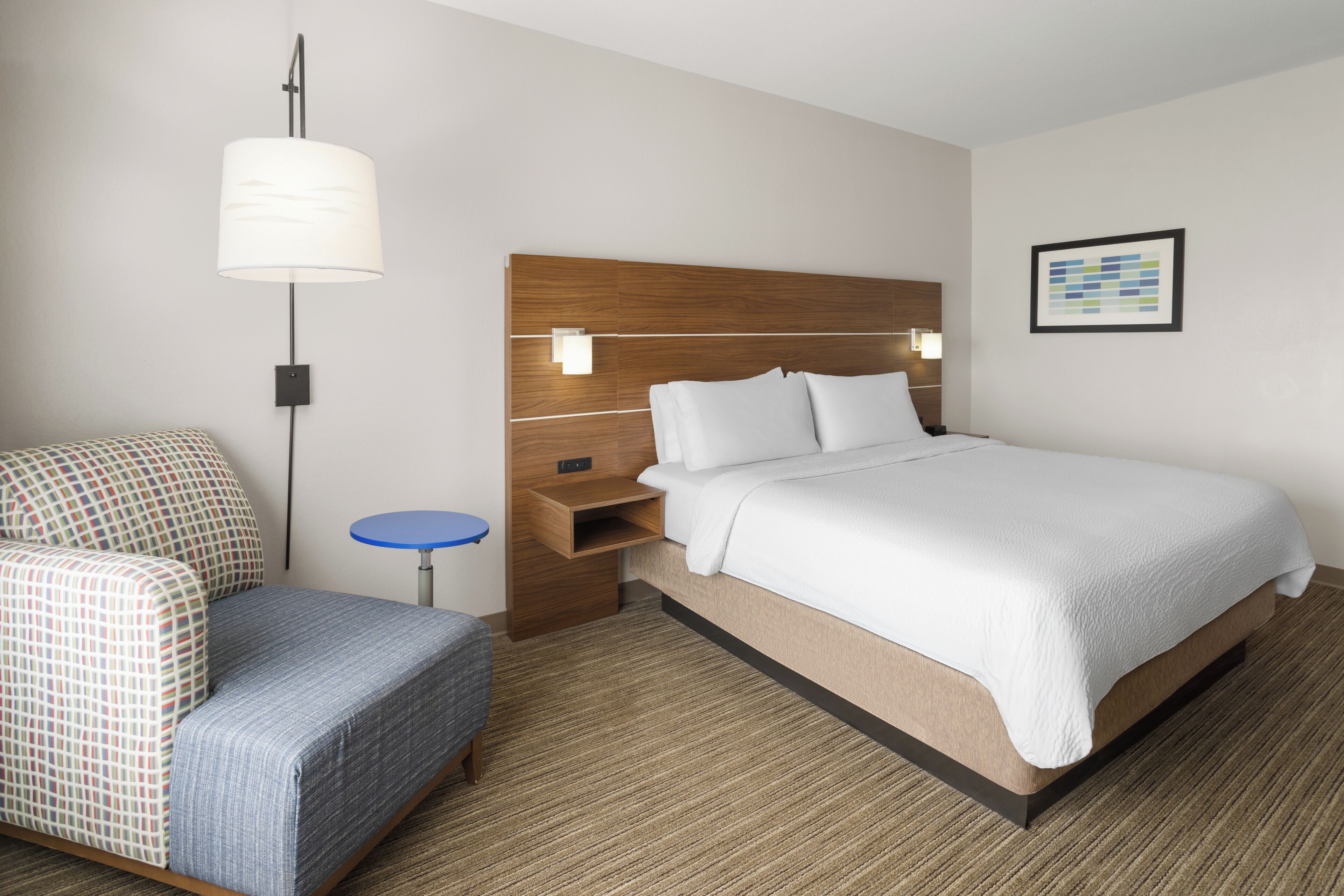 A hotel room with a bed, a chair, a small table, a wall-mounted lamp, and a framed picture.