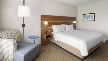 A hotel room with a bed, a chair, a small table, a wall-mounted lamp, and a framed picture.