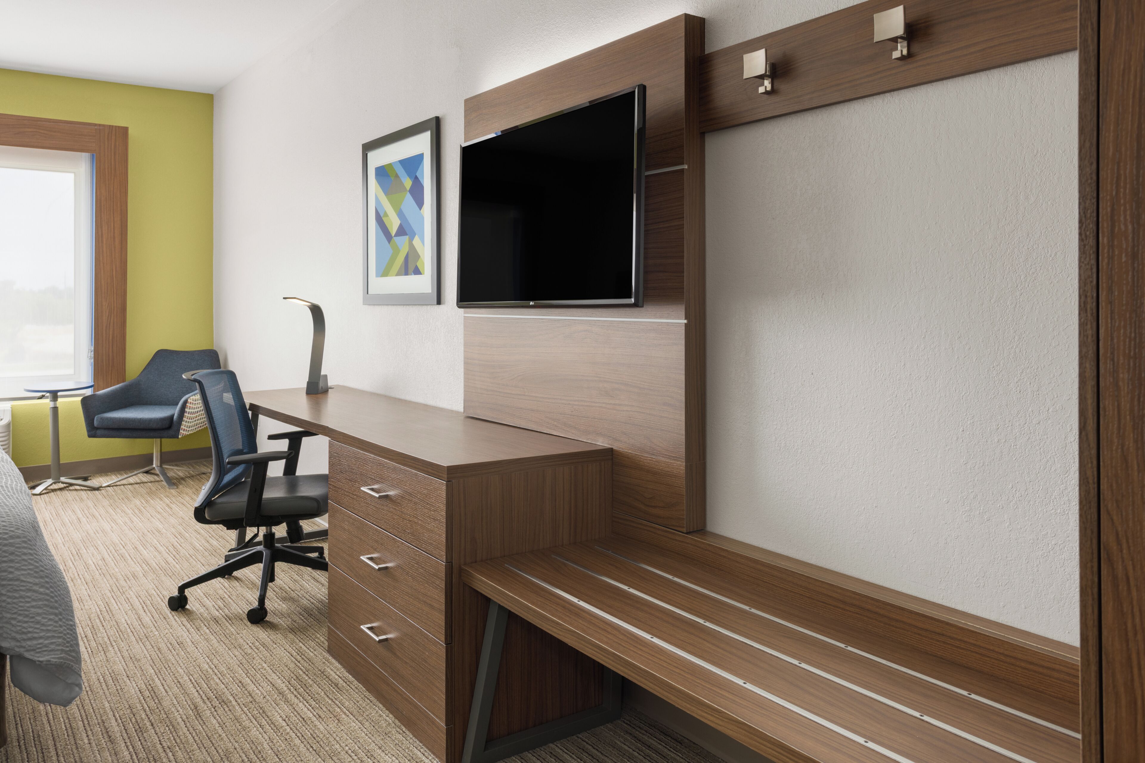 A hotel room with a wooden desk, a flat-screen TV, a blue office chair, and a bed with a gray blanket.