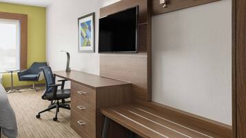 A hotel room with a wooden desk, a flat-screen TV, a blue office chair, and a bed with a gray blanket.