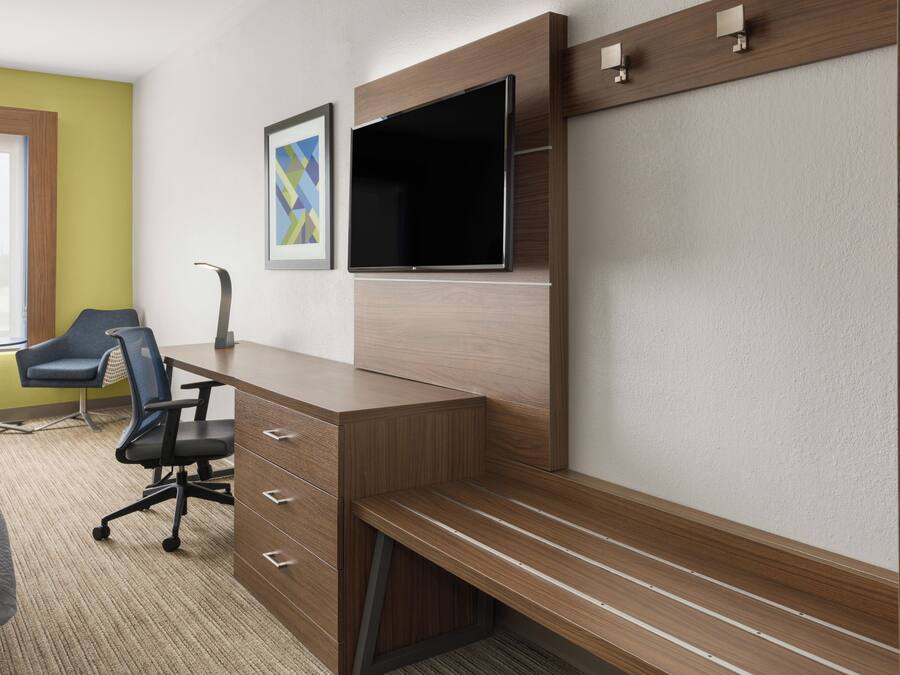 A hotel room with a wooden desk, a flat-screen TV, a blue office chair, and a bed with a gray blanket.
