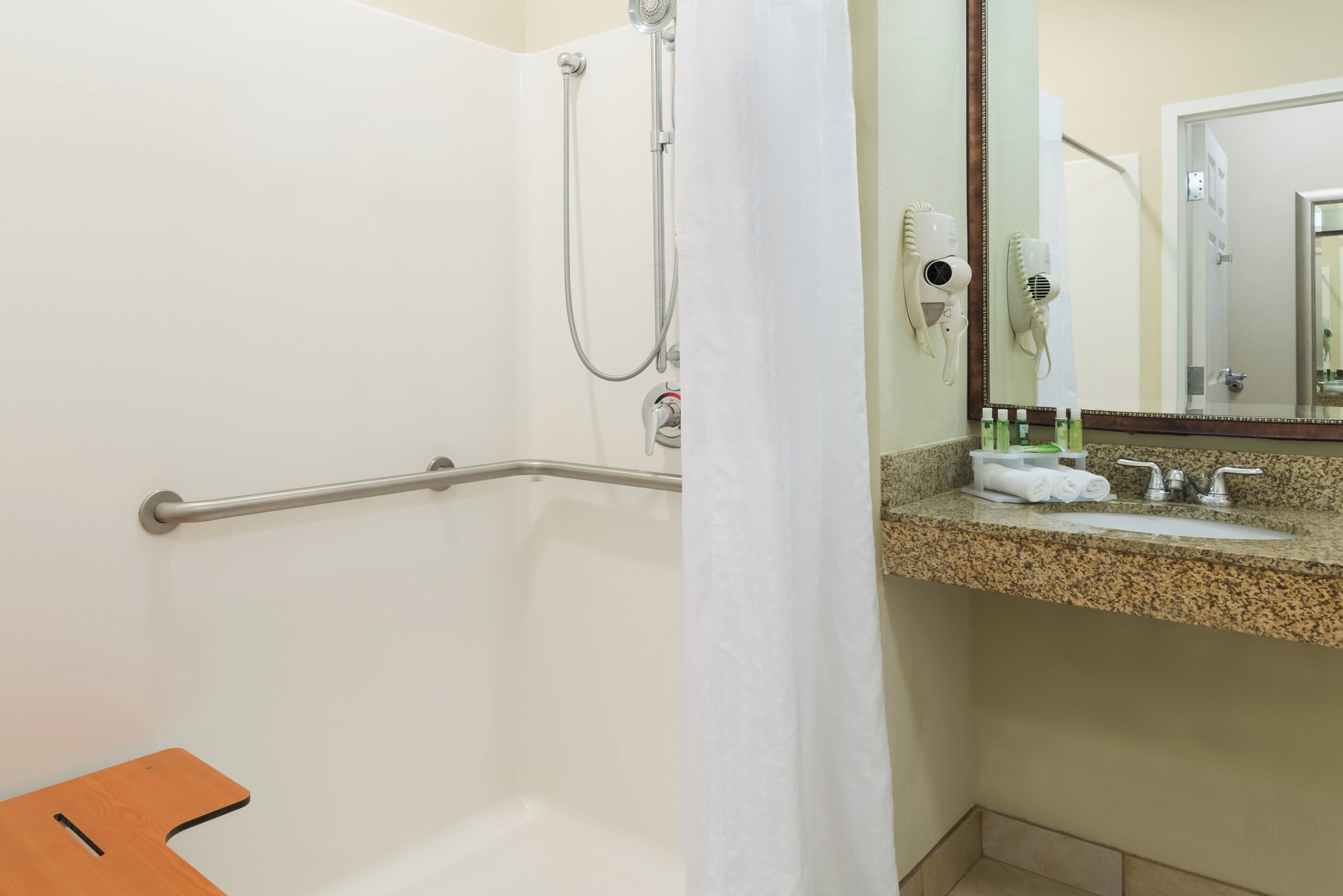 A bathroom with a shower, a handrail, a sink with toiletries, and a mirror.