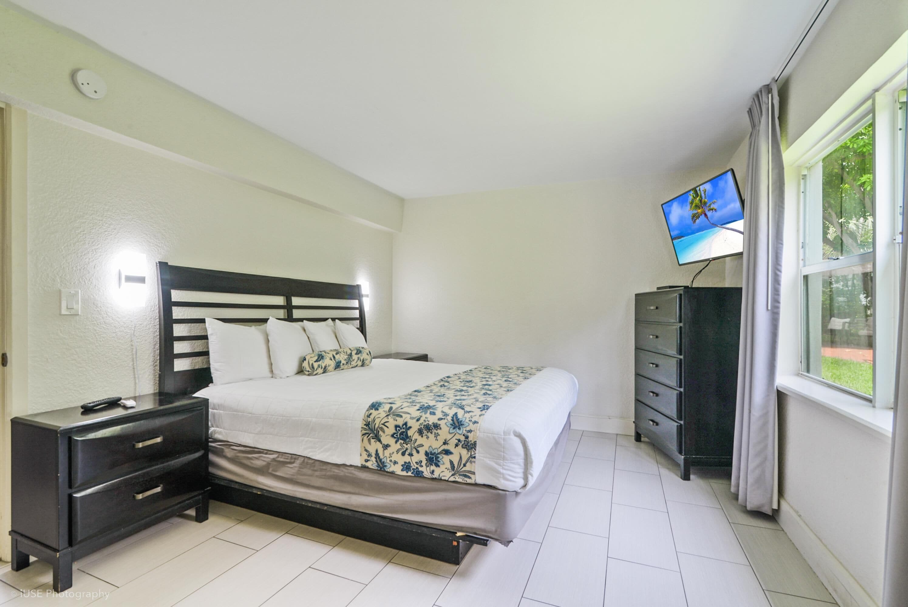 A neatly arranged bedroom with a bed, a nightstand, a dresser, and a television mounted on the wall.
