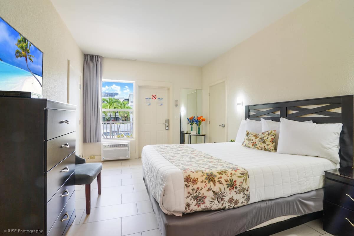 A hotel room with a bed, a dresser, a TV, and a balcony with a view of the beach.