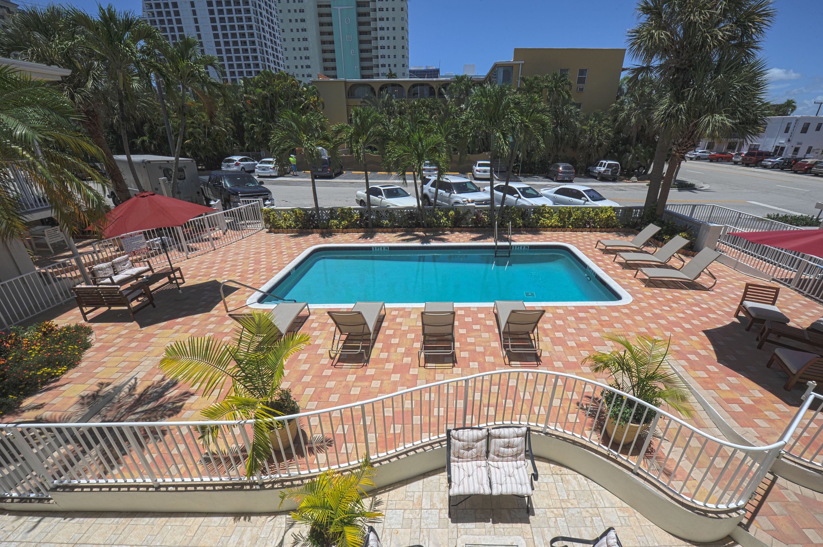 A swimming pool with lounge chairs and a red umbrella, surrounded by palm trees and a brick patio.