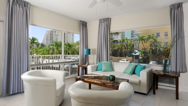 A spacious living room with a white sofa, two armchairs, a coffee table, and a balcony with a view of palm trees and buildings. - Soleado Hotel (Fort Lauderdale)