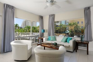 A spacious living room with a white sofa, two armchairs, a coffee table, and a balcony with a view of palm trees and buildings. - Soleado Hotel (Fort Lauderdale)