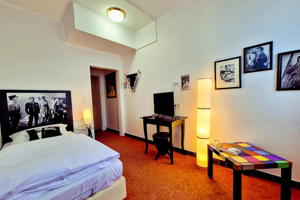 Quarto de hotel com cama, escrivaninha, luminária e quadros na parede.