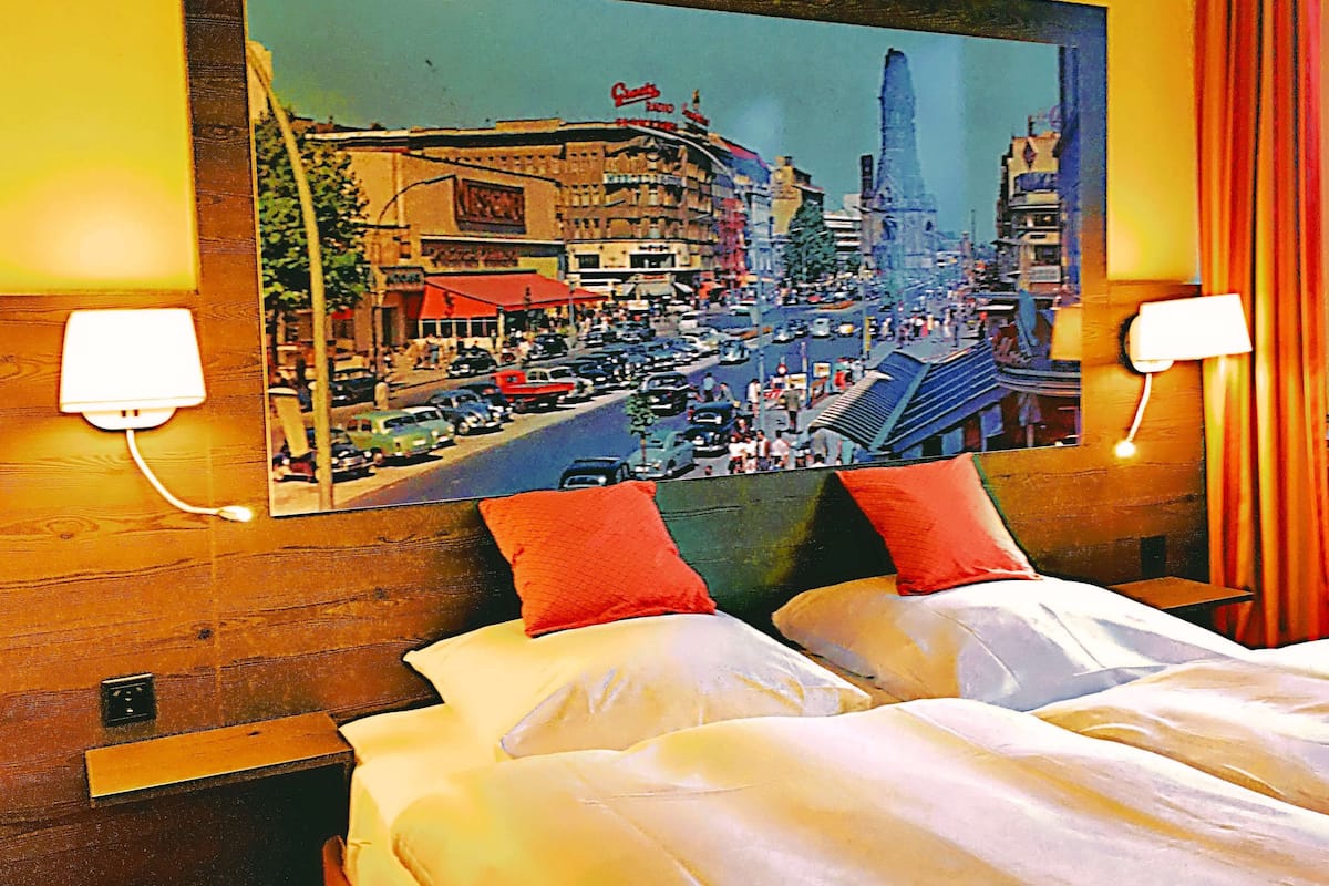 Quarto de hotel com uma grande pintura da paisagem urbana na parede, duas camas brancas com almofadas vermelhas e duas luminárias de cabeceira.