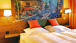 Une chambre d’hôtel avec un grand tableau représentant un paysage urbain au mur, deux lits blancs avec des oreillers rouges et deux lampes de chevet.