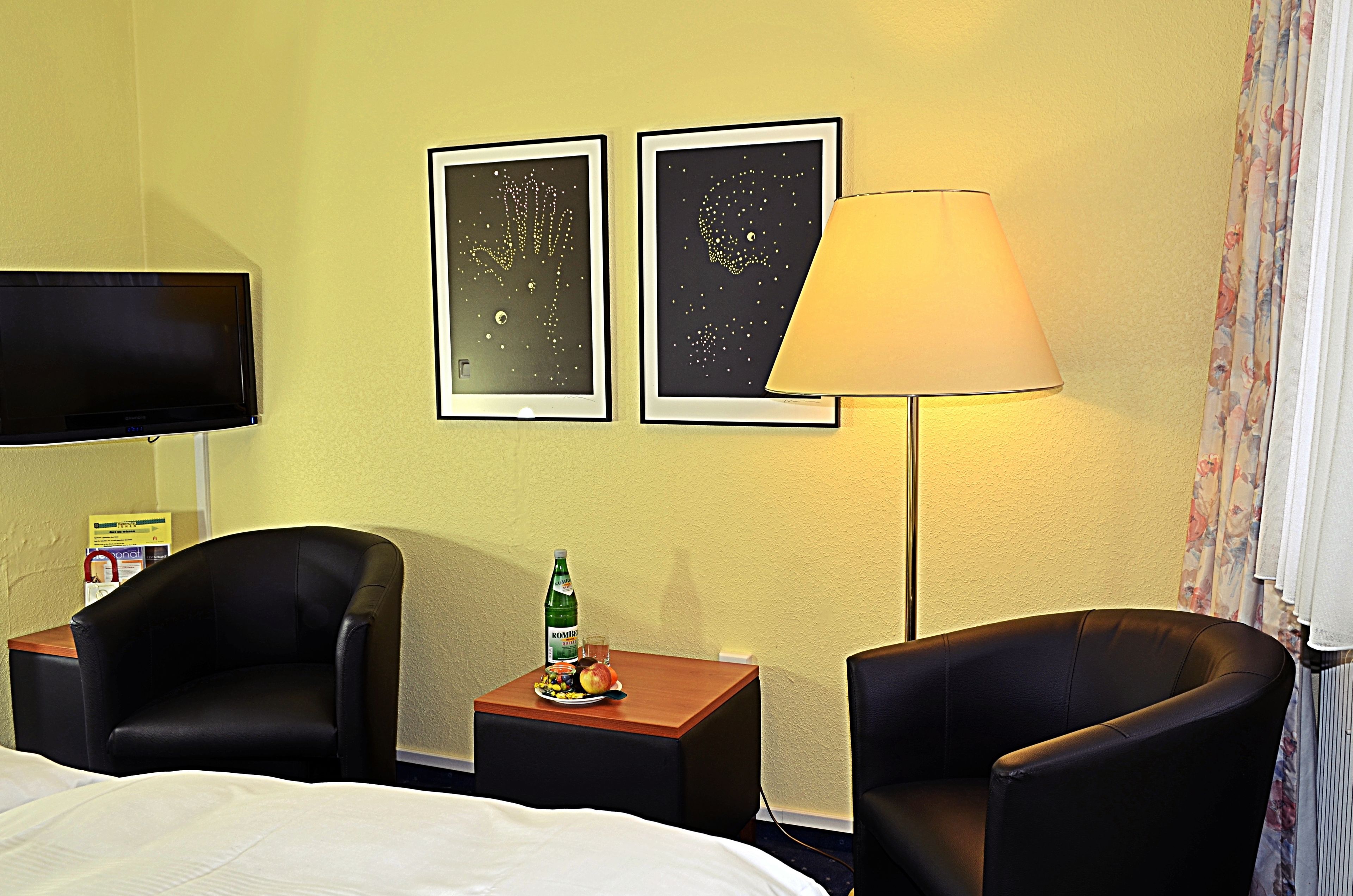 Ein Hotelzimmer mit einem Bett, zwei Sesseln, einem kleinen Tisch mit einer Flasche und Obst, einem Fernseher und zwei gerahmten Bildern an der Wand.