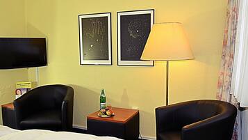 Ein Hotelzimmer mit einem Bett, zwei Sesseln, einem kleinen Tisch mit einer Flasche und Obst, einem Fernseher und zwei gerahmten Bildern an der Wand.
