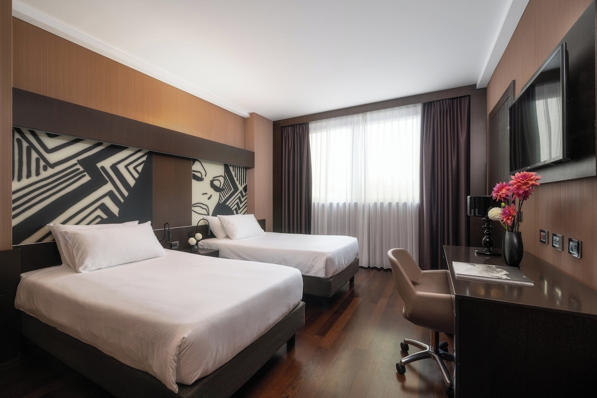 Une chambre d’hôtel avec deux lits, un bureau, une chaise, une télévision et une grande fenêtre avec des rideaux.