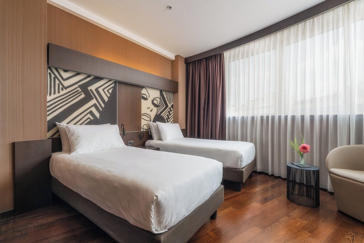 Une chambre d’hôtel avec deux lits, une petite table ronde ornée d’un vase et une chaise.