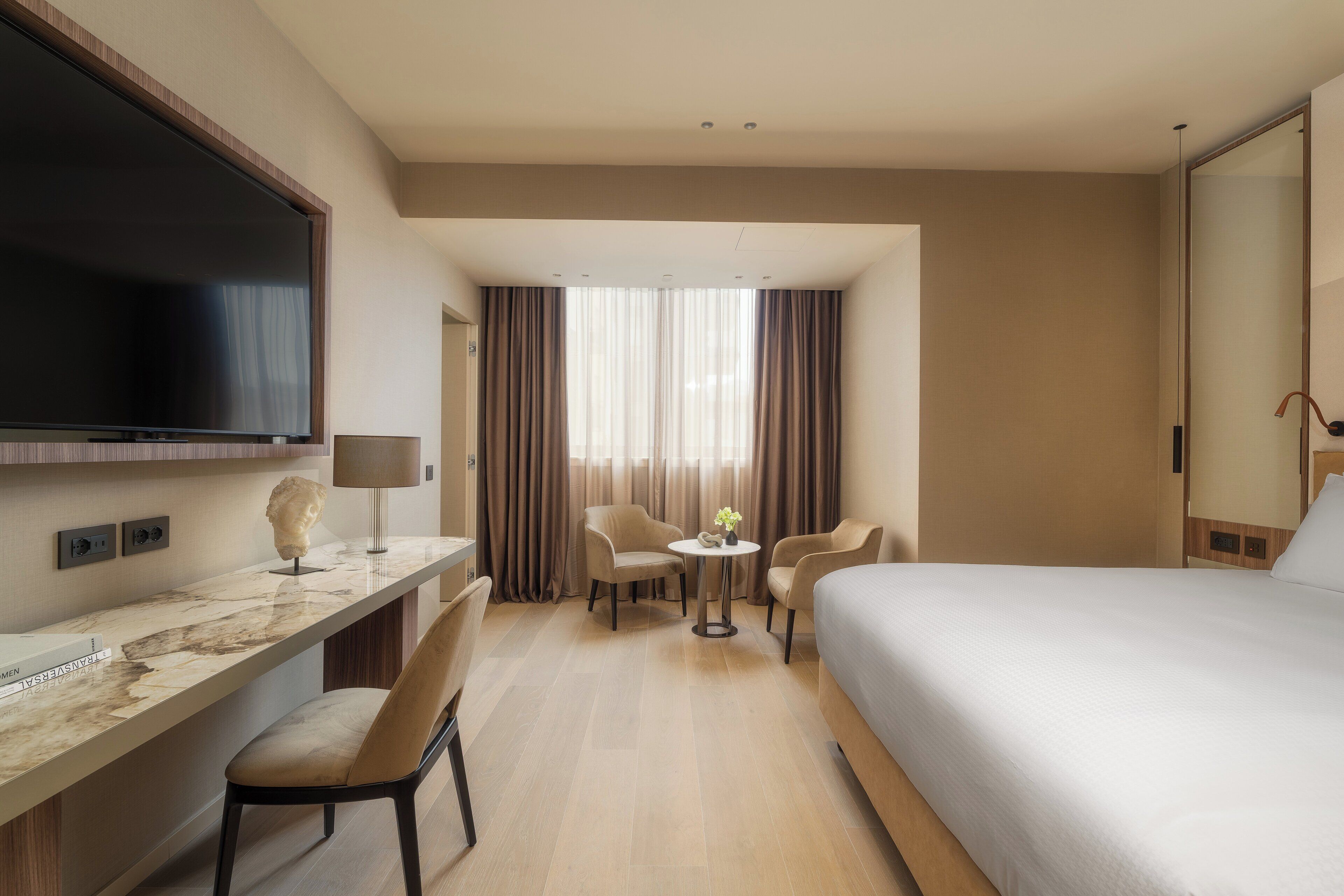 A hotel room with a bed, a TV, a desk with a chair, a small table with two chairs, and a window with curtains.