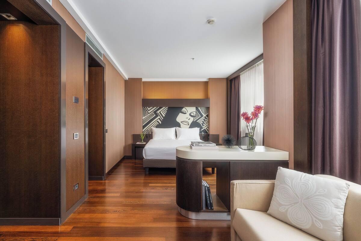 Una habitación de hotel con cama, escritorio, silla y un amplio ventanal con cortinas.