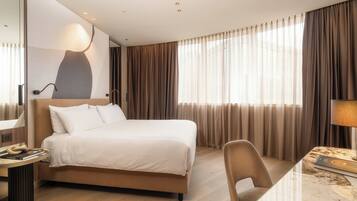 Habitación de hotel con cama, escritorio, silla, lámpara y un ventanal con cortinas.