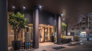 A modern hotel entrance with a glass door, potted plants, and a covered walkway.