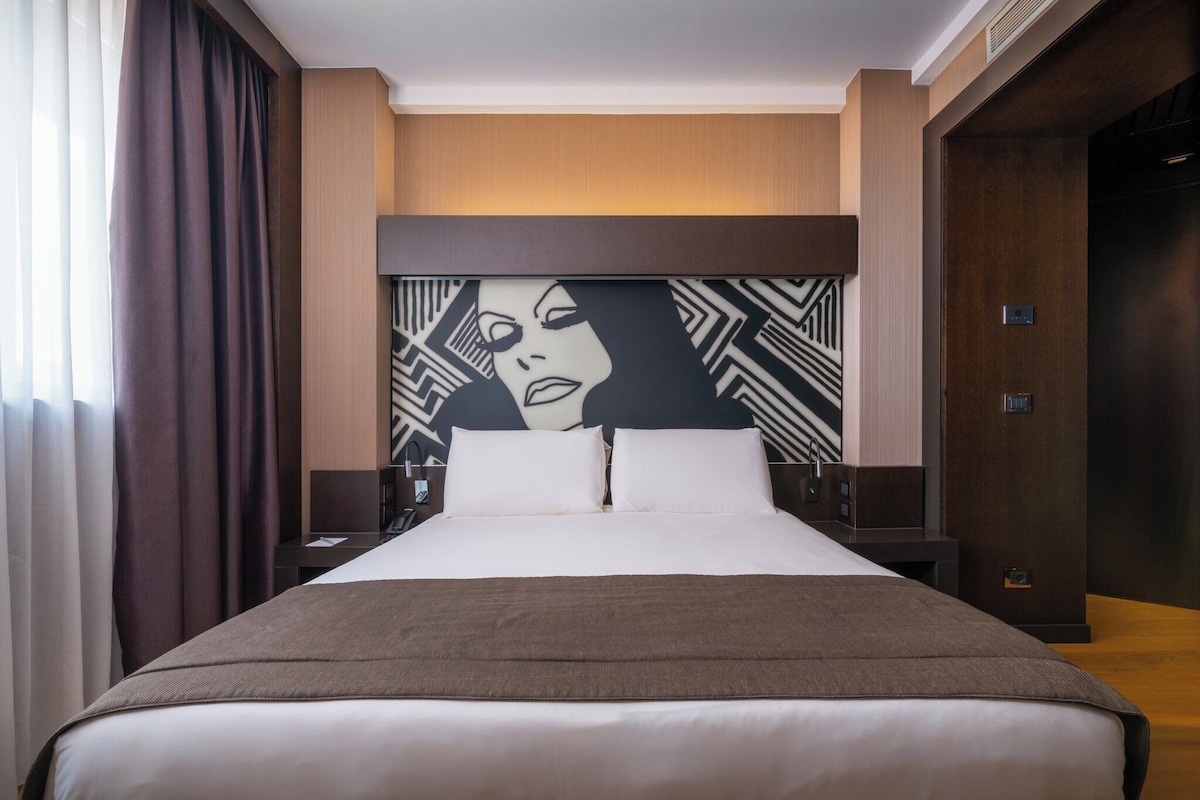Une chambre d’hôtel avec un grand lit, une tête de lit ornée d’un motif géométrique, une table de chevet et une porte en bois.