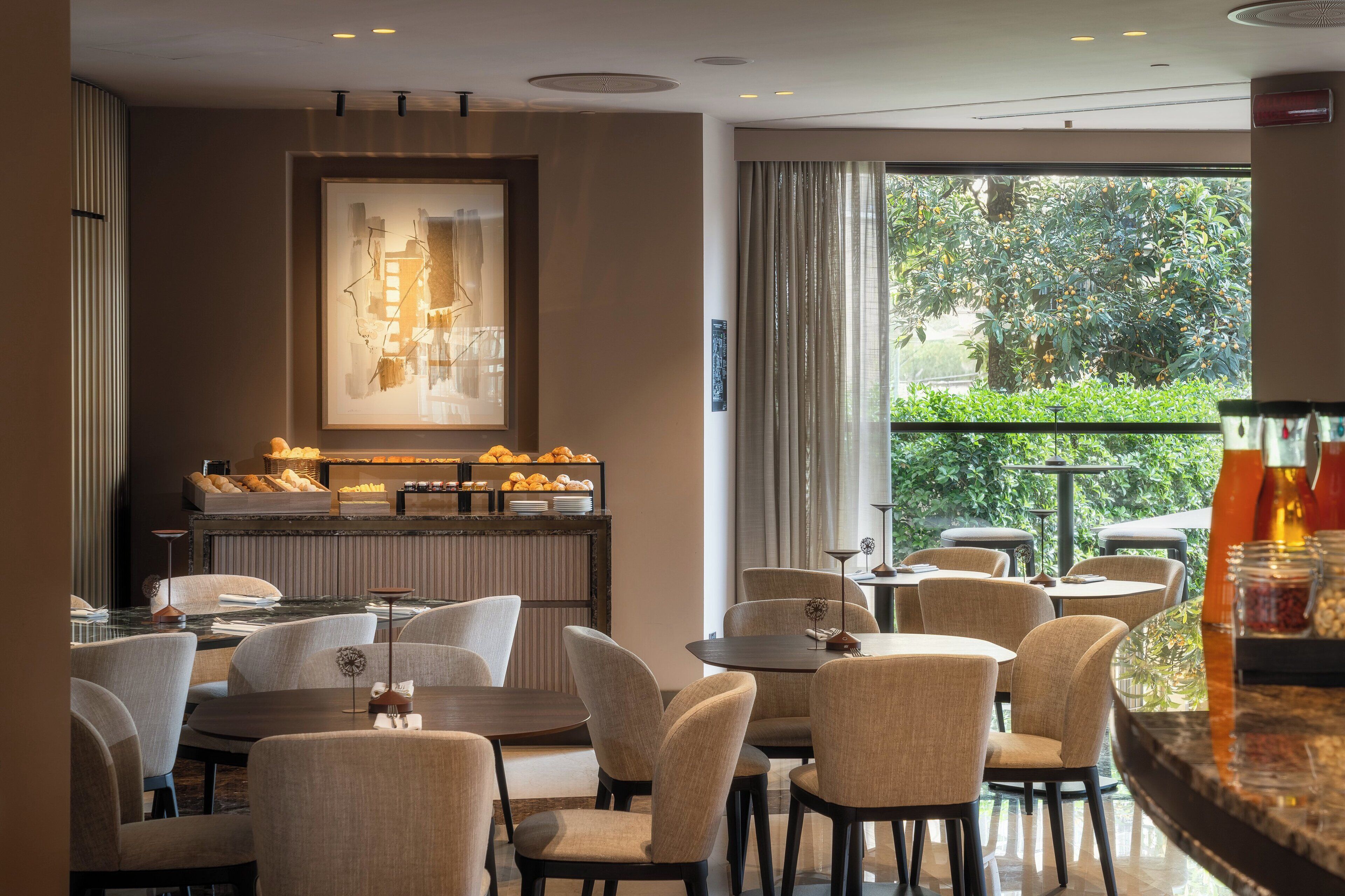 A modern dining area with round tables and chairs, a fruit display, and a view of greenery through large windows.