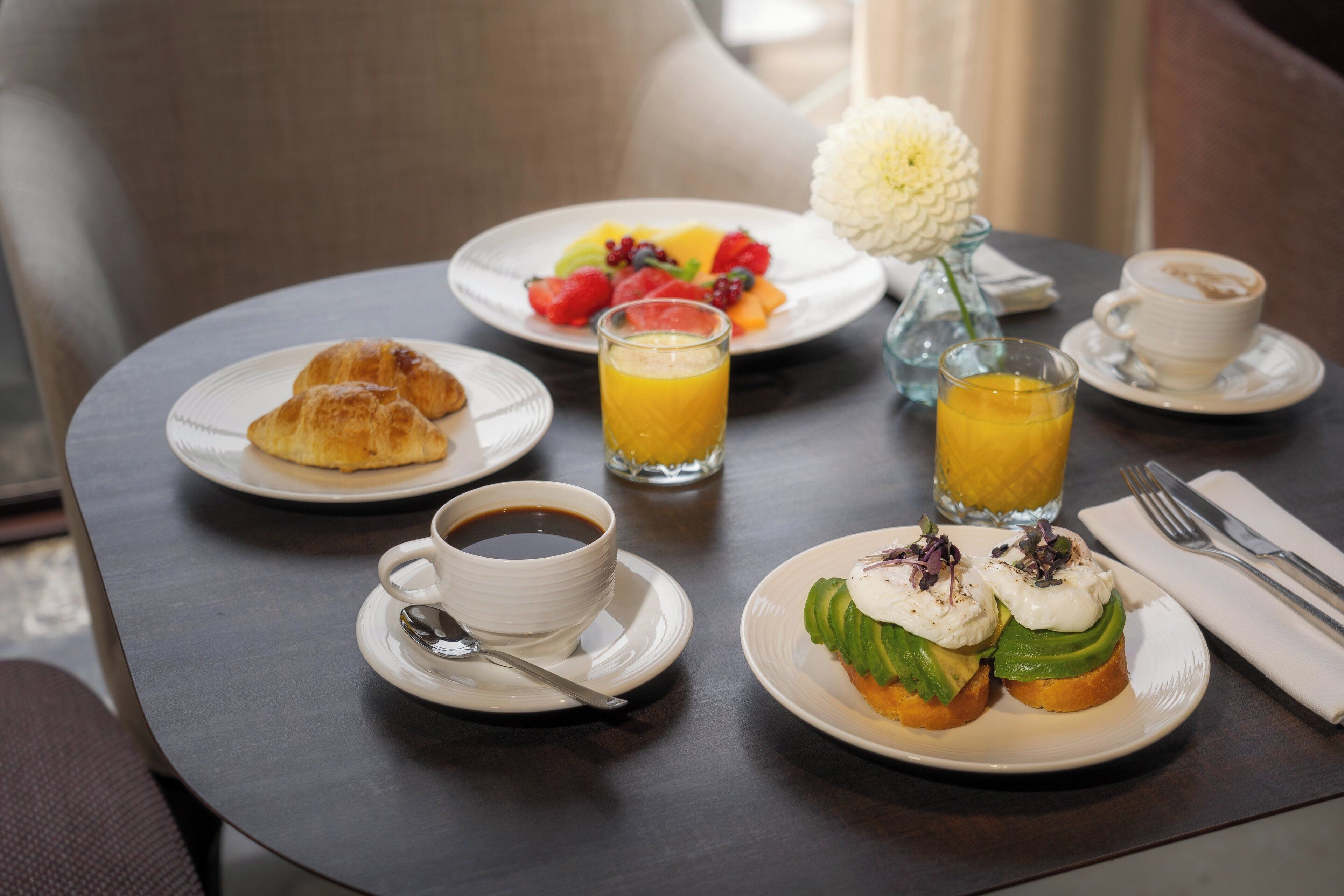 A breakfast setting with croissants, fruit, avocado toast, coffee, and orange juice on a table.