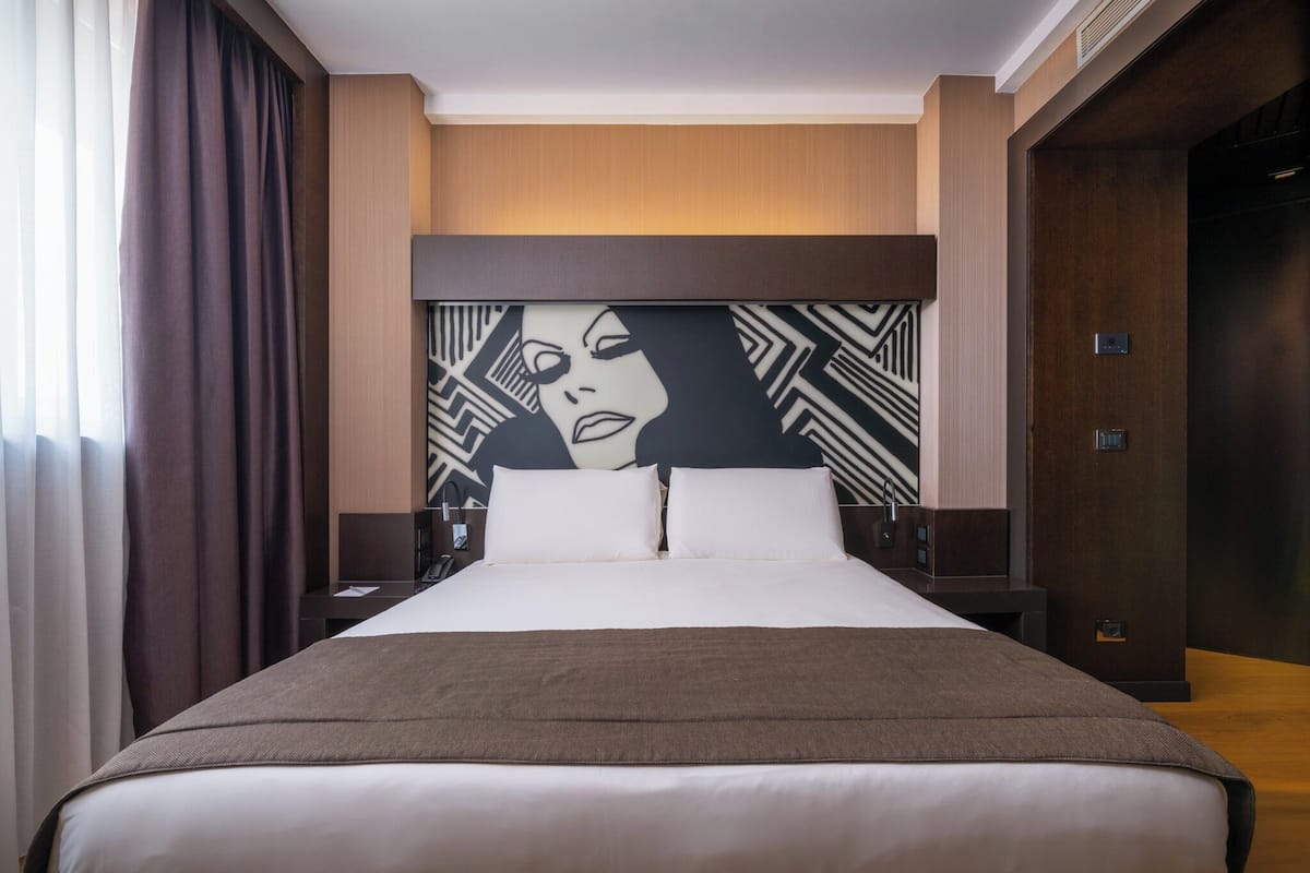 Habitación de hotel con una cama grande, un cabecero con diseño geométrico, una mesita de noche y una puerta de madera.