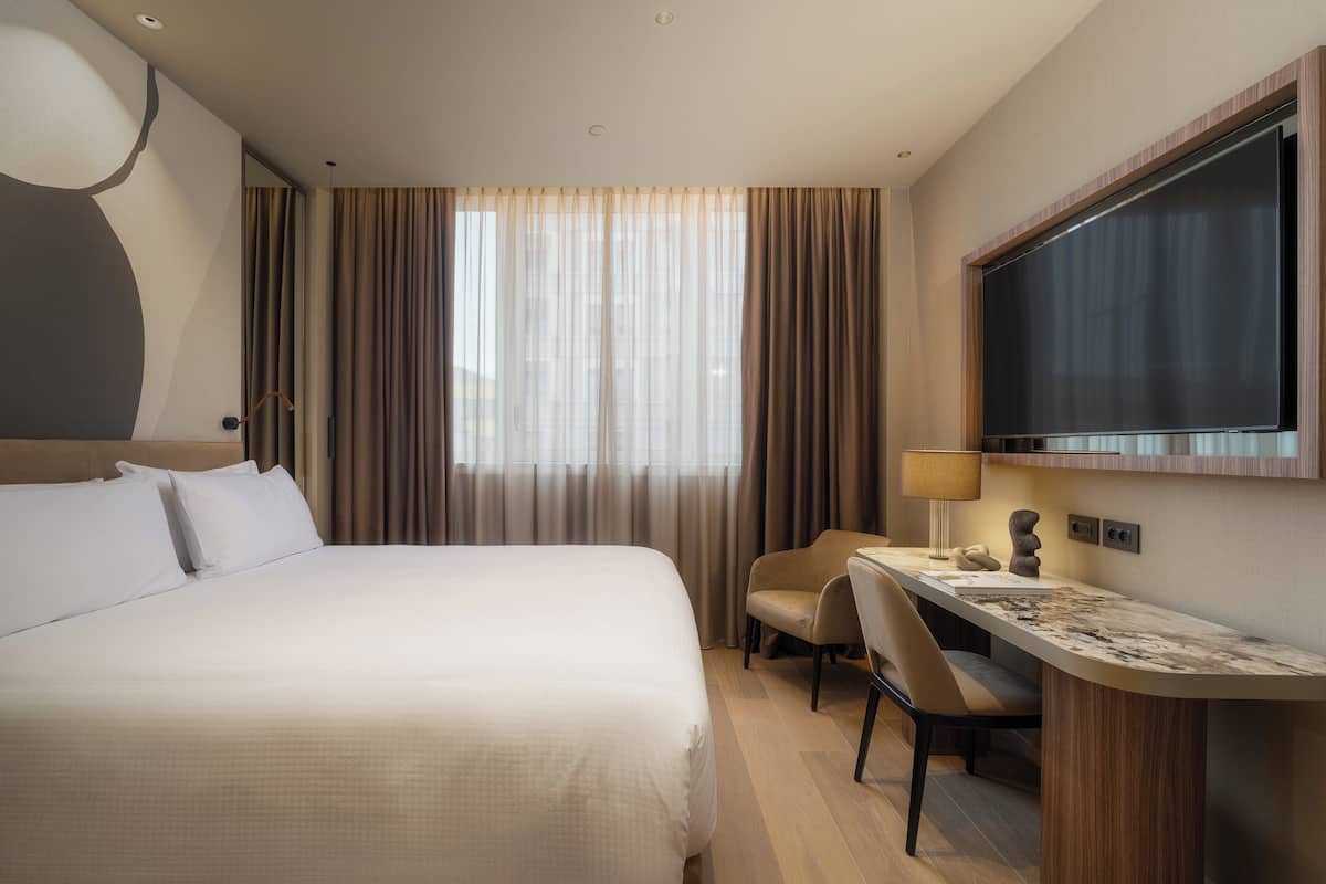 Una habitación de hotel con cama, escritorio, silla, televisión y ventana con cortinas.