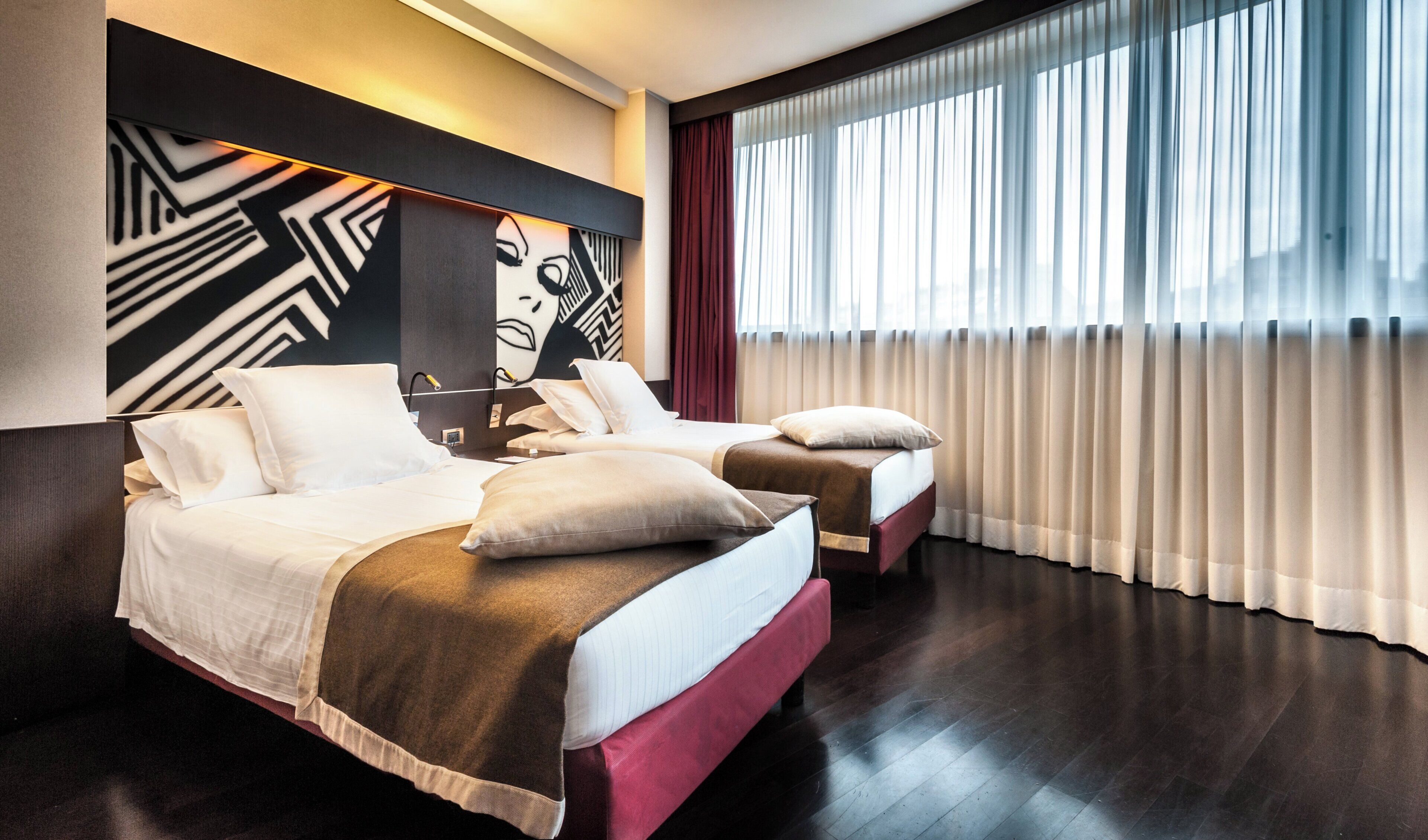 Ein Hotelzimmer mit zwei Betten, einem großen Fenster mit durchsichtigen Vorhängen und einem modernen Kunstwerk an der Wand.