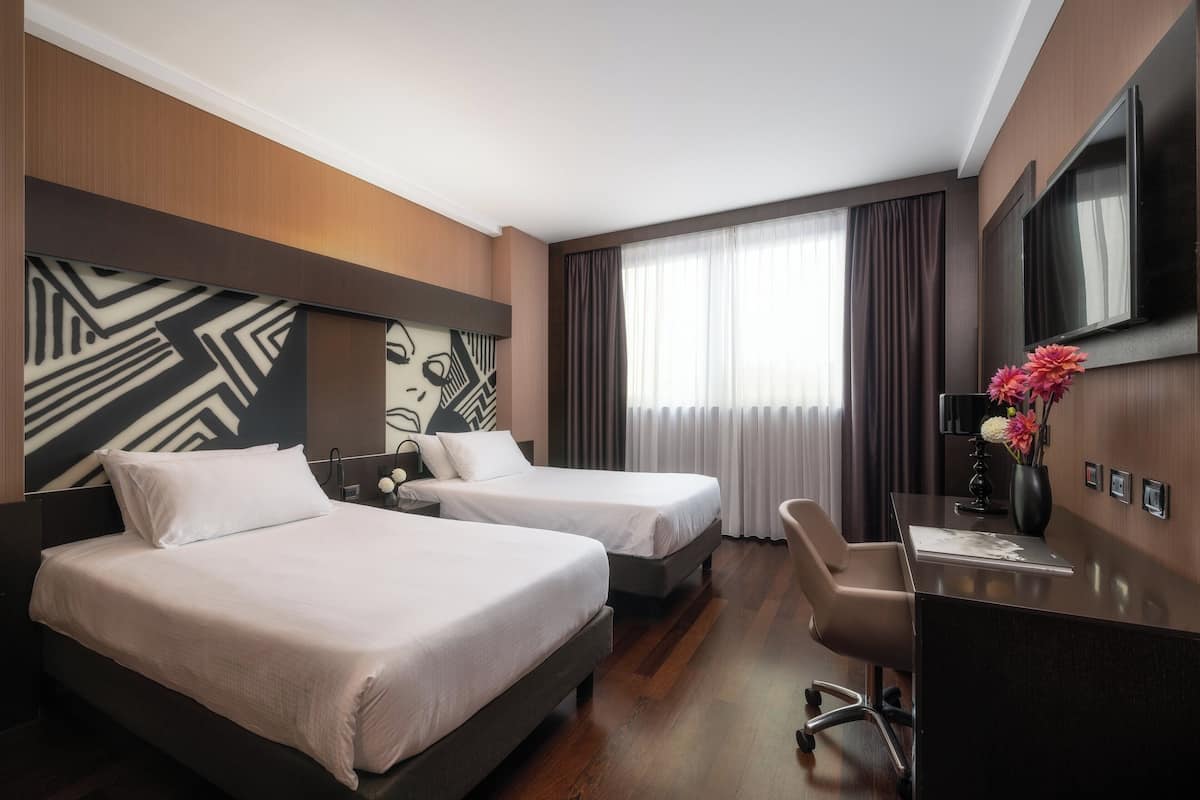 Habitación de hotel con dos camas, un escritorio, una silla, un televisor y un ventanal con cortinas.