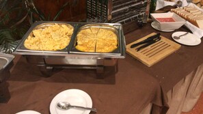 A buffet setup with two large metal trays of food, a wooden cutting board with a knife, and a white plate with a spoon. - Rural Hotel Las Gacelas (Becerril de la Sierra)