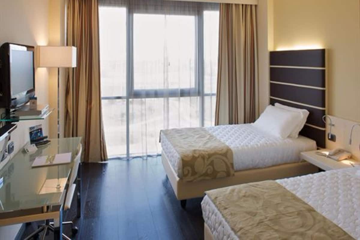 Habitación de hotel con dos camas, un escritorio con ordenador, televisor y un ventanal con cortinas.
