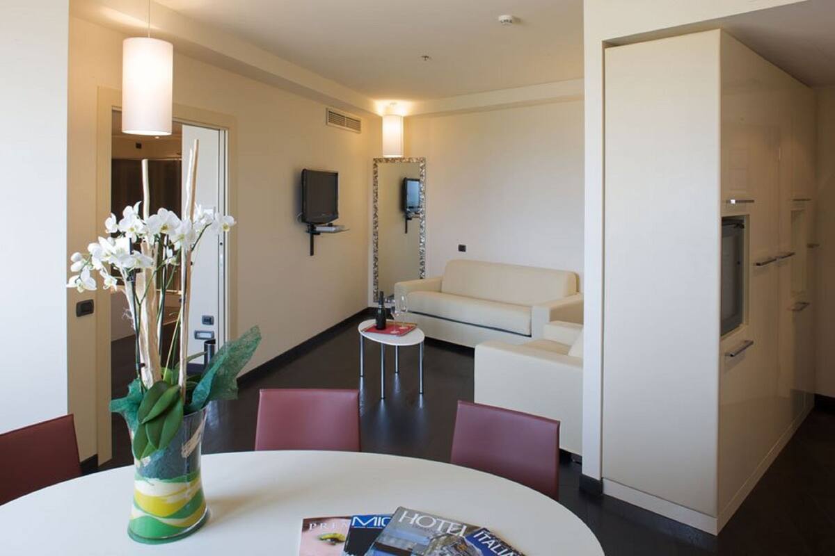 Una habitación de hotel moderna con un sofá blanco, una mesa de comedor con sillas, un televisor montado en la pared y un minibar.