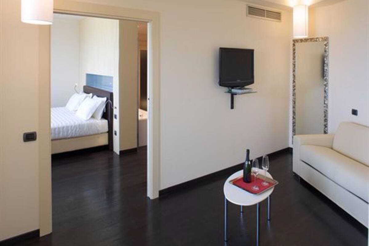 Una habitación de hotel con una cama, un televisor, una mesita con una botella y un vaso, y un sofá blanco.