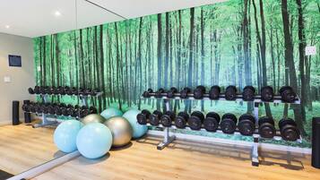 Une salle de sport ornée d’une fresque murale représentant une forêt, avec des rangées d’haltères et des ballons d’exercice.