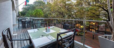 An outdoor dining area with tables set for two, surrounded by a wooden deck and metal railings.