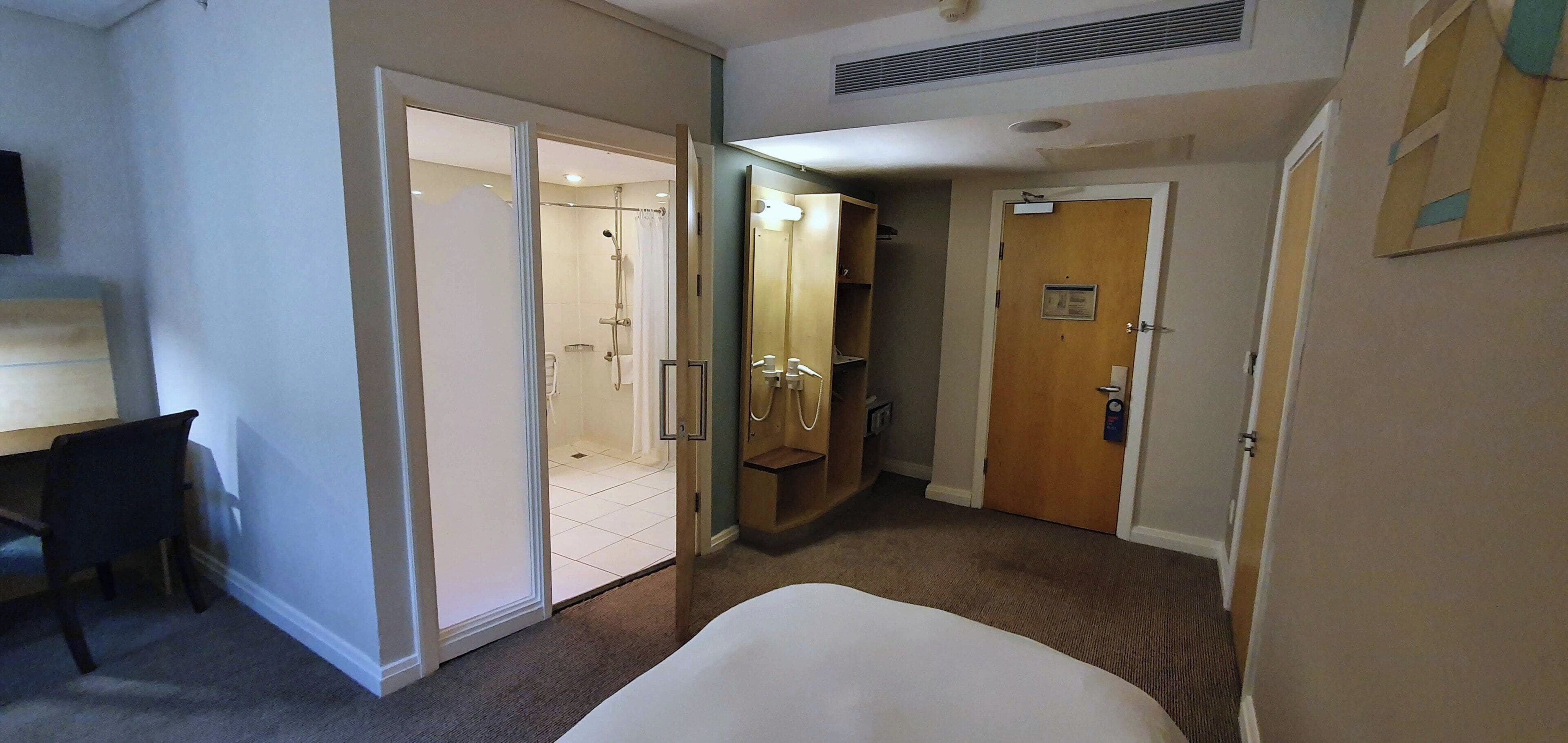 A hotel room with a bed, a desk, a shower area, and a door leading to another room.