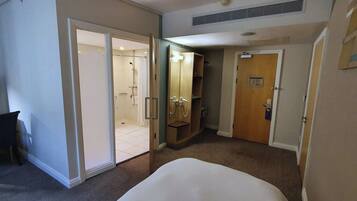 A hotel room with a bed, a desk, a shower area, and a door leading to another room.