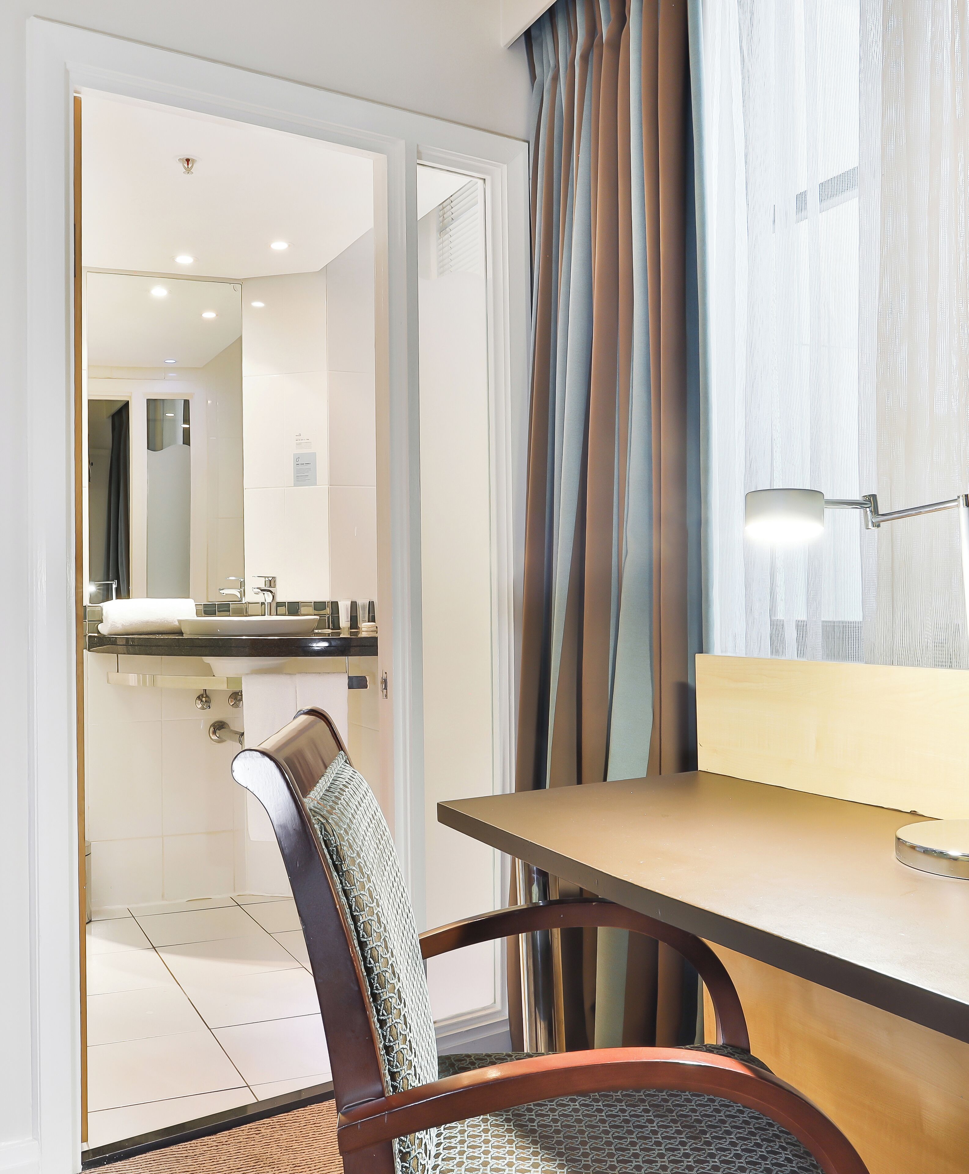 Habitación de hotel con escritorio de madera, una silla, un espejo y un baño visible a través de una puerta abierta.