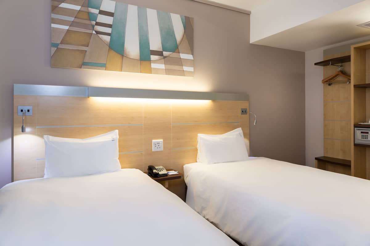 A hotel room with two beds, a wooden headboard, a wall-mounted artwork, a bedside table with a phone, and a wooden wardrobe.