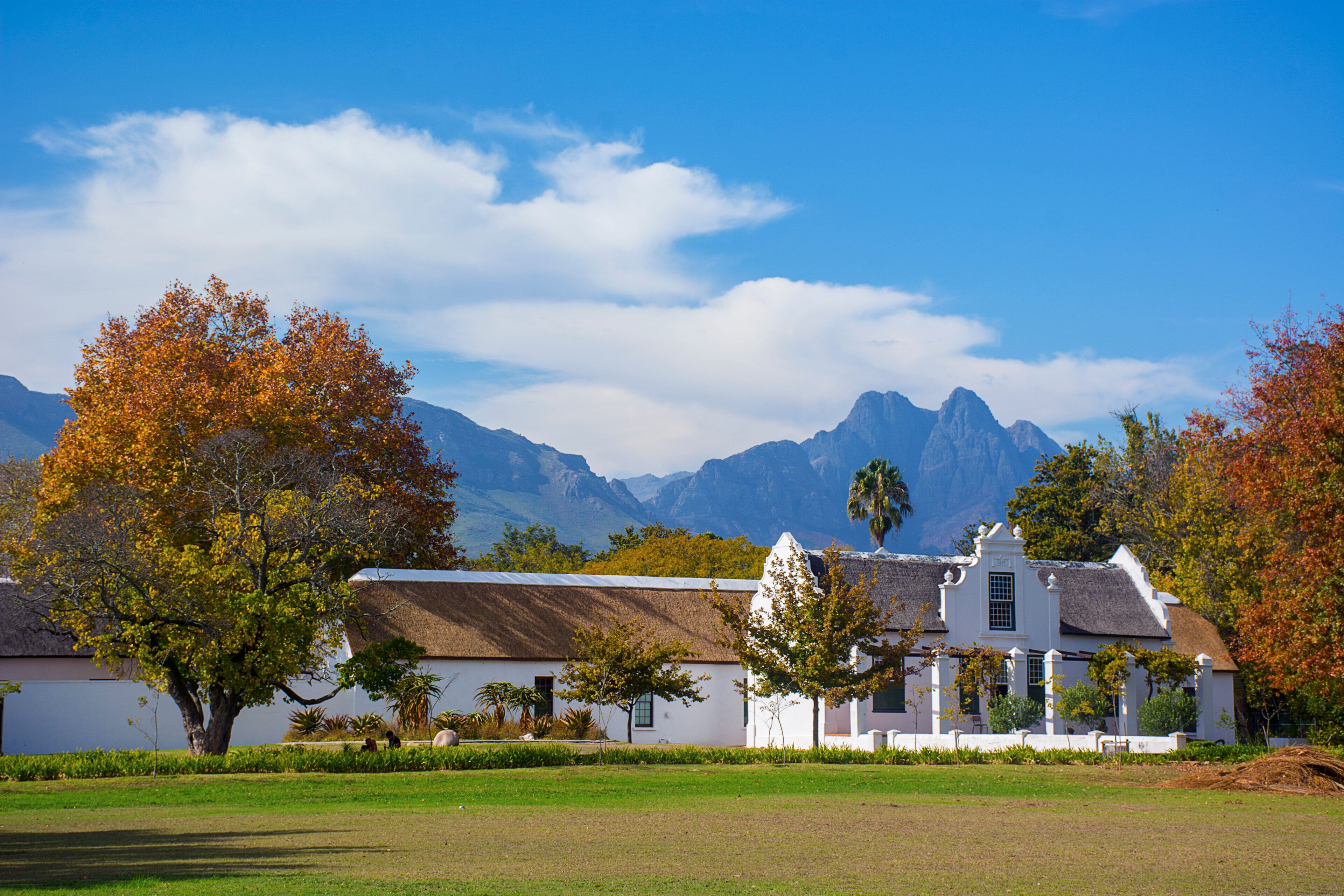 A white building with a thatched roof, surrounded by trees and a grassy field, with mountains in the background.