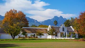 A white building with a thatched roof, surrounded by trees and a grassy field, with mountains in the background.