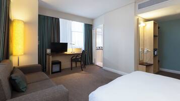 A hotel room with a bed, a desk with a television, a chair, a sofa, and a bathroom with a mirror and a telephone.
