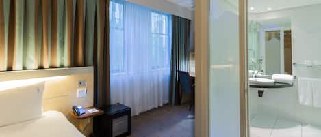 A hotel room with a bed, bedside table, phone, and a view of the bathroom through a glass door.
