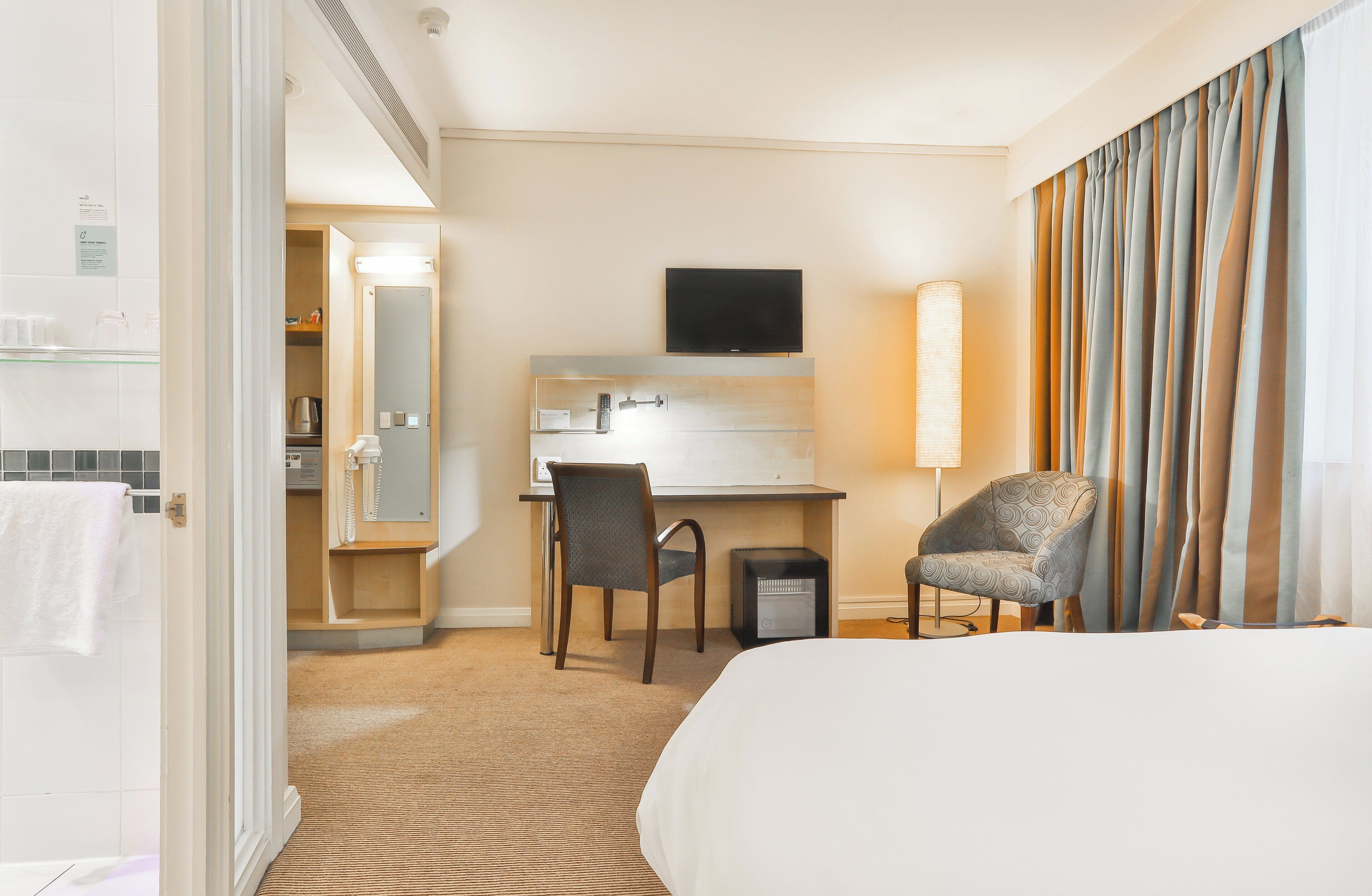 A hotel room with a bed, a desk with a chair, a television, and a large window with curtains.
