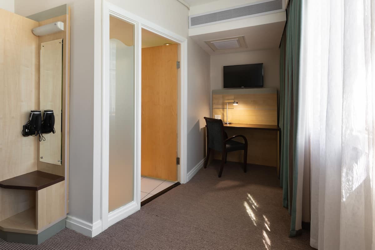 A hotel room with a wooden door, a desk with a chair, a flat-screen TV, and a telephone.