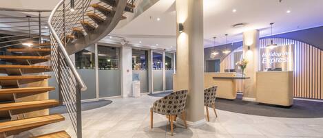 Un hall d’hôtel moderne avec un escalier en colimaçon, un comptoir d’accueil et deux fauteuils à motifs.